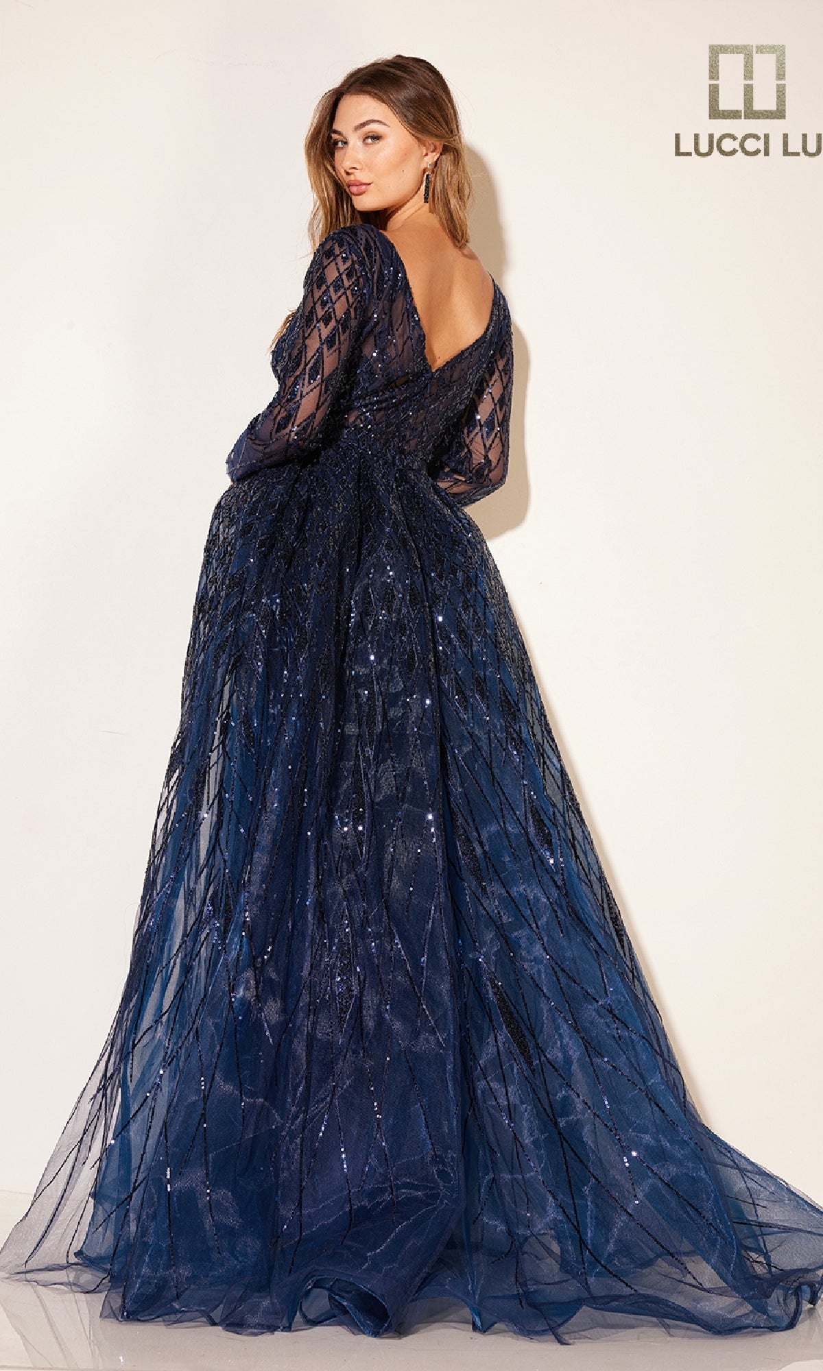 A woman poses against a light background in the Lucci Lu C8095 long formal dress, featuring dark blue sequins, sheer sleeves, an open back, and a hint of tulle overskirt.